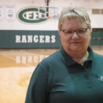 -middle-aged woman with short hair and glasses with a green FHC logoed polo shirt on stands in a high school gym at Forest Hills Central High School