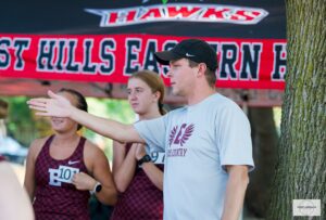 Man with a black baseball cap standing in from of an FHE tent talks to high school runners