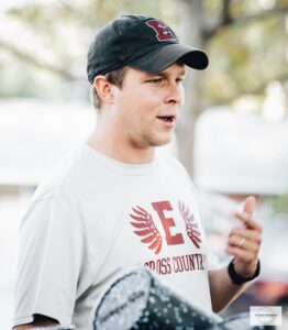 A man with a baseball hat and a tshirt with an E on it talks to high school athletes outside