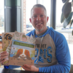 A man sitting in a chair holding up three children's books.