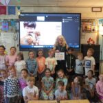 a group of students stand in front of a large tv screen with a student's face on the tv screen