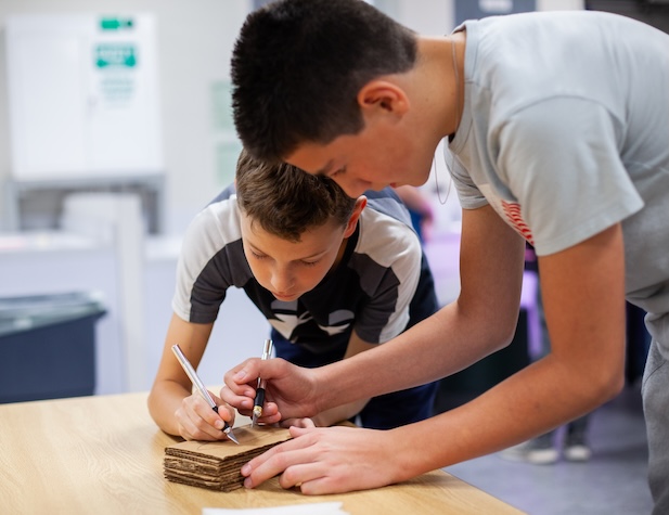 two male students cutting out shapes out of cardboard with exacto knives