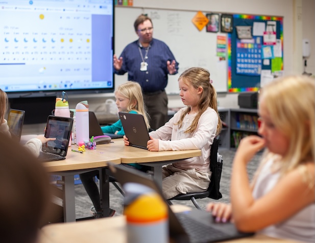 young female students working on computers in class while the teacher is talking